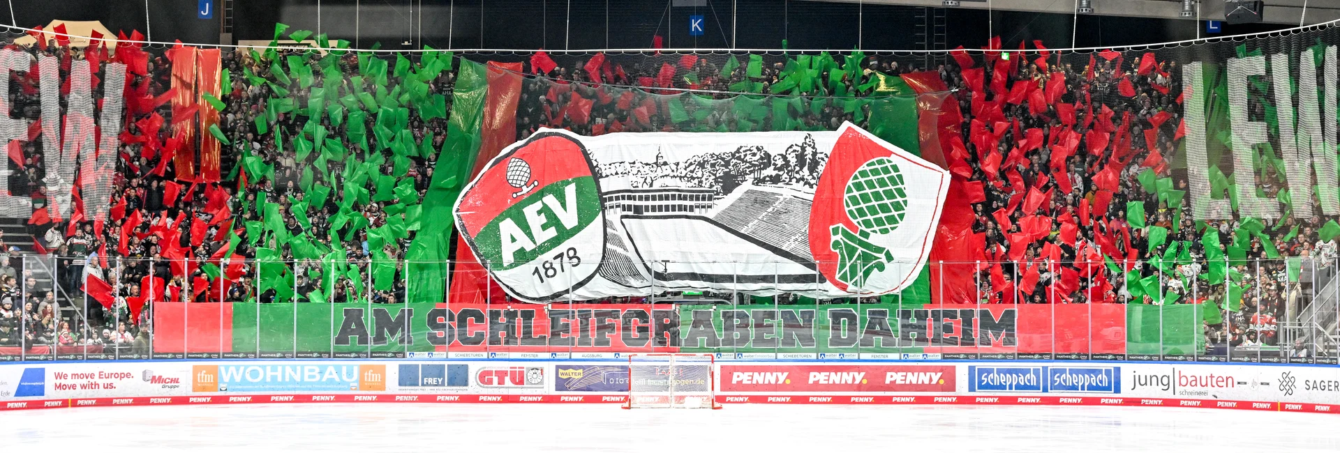 Fans halten Rote und grüne Flaggen hoch. Das Gesamtbild ergibt ein Rot-Grün-Muster. In der Mitte ist das AEV-Logo, das historische Curt-Frenzel-Stadion sowie das Augsburger Wappen. Darunter steht groß: "Am Schleifgraben daheim"