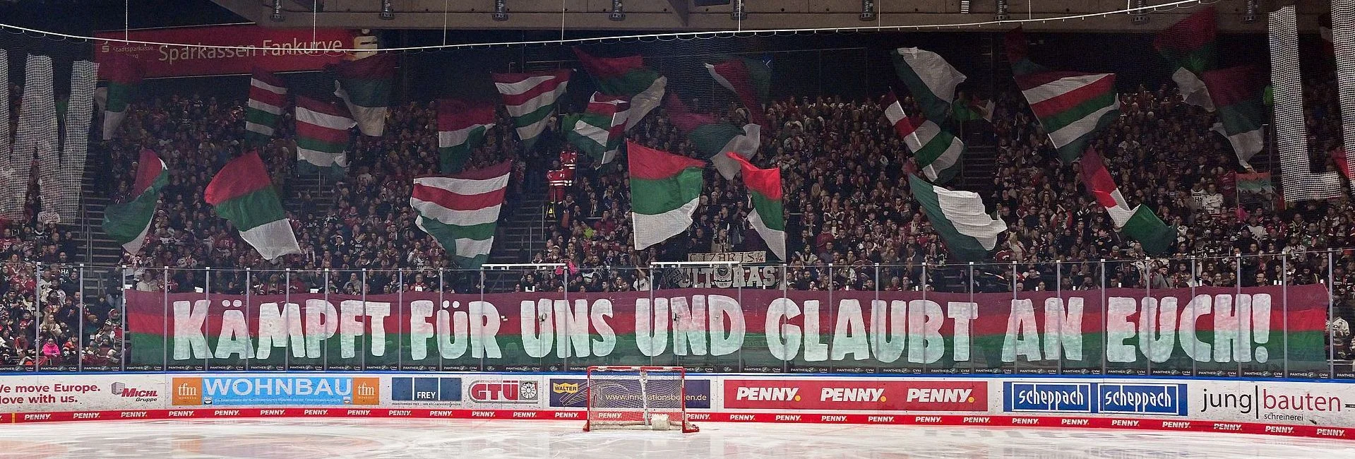 Blick in die Fankurve, die einen großen Schriftzug "Kämpft für uns und glaubt an euch!" hochhalten. Es werden viele, große AEV-Flaggen geschwenkt.